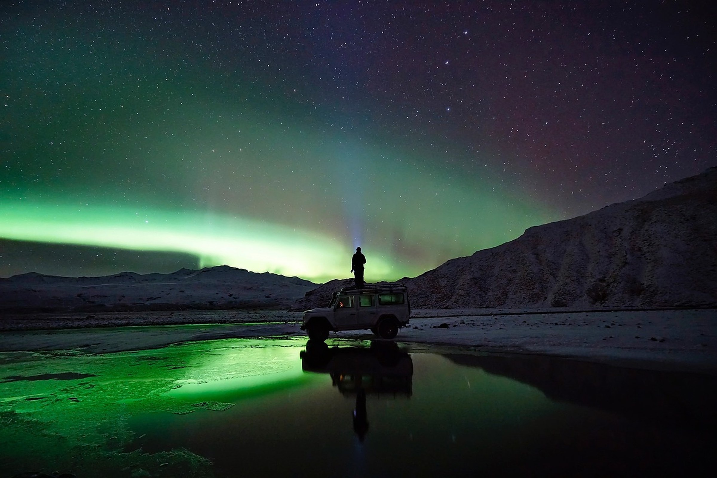 A person standing on the roof of a 4×4 under the green northern lights